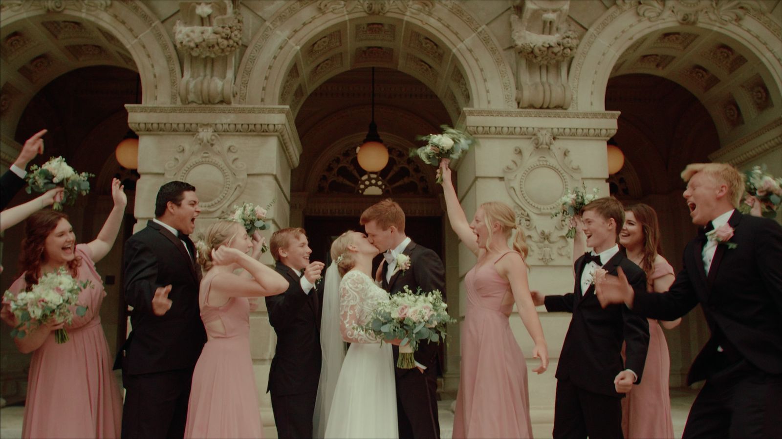 Couple kissing under stone arches as their wedding party cheers
